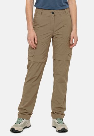 Persoon draagt bruine om te zetten cargobroek opengehouden bij de knieën en beige wandelschoenen met groene veters, staand tegen een witte achtergrond.