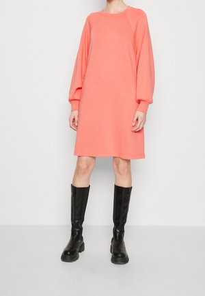 Kobieta ubrana w koralową sukienkę na długi rękaw, sięgającą kolan, z bufkami na rękawach oraz czarne, masywne kozaki za kolano na tle jednolitego tła.