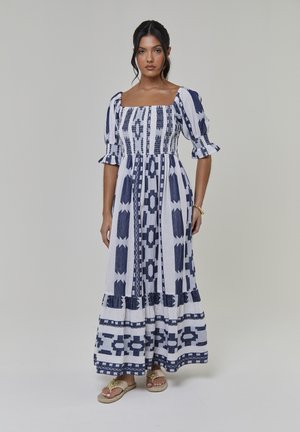 Robe longue blanche avec des motifs géométriques bleu marine. Elle présente un col carré, des manches bouffantes, un corsage froncé et un ourlet à volants à paliers.