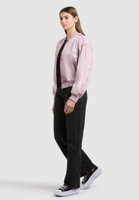 Femme debout portant une veste bomber violette claire, un t-shirt noir, un jean noir et des baskets noir et blanc, sur un fond clair uni.