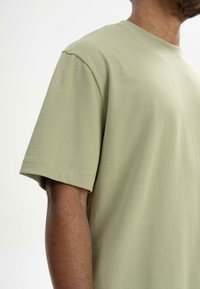 Hellgrünes T-Shirt mit kurzen Ärmeln aus weicher Baumwolle, das einen lockeren Schnitt und einen genähten Saum aufweist, mit einer glatten Textur und ohne sichtbare Muster.
