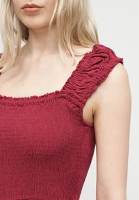 Szőke nő, aki egy ujjatlan, mélyvörös, texturált felsőt visel, széles fodros pántokkal és négyzet alakú nyakkivágással, egyszínű háttér előtt.