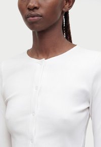 Gros plan d'une personne portant une chemise blanche côtelée à manches longues avec un col rond et de petits boutons sur le devant.