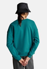Felpa teal con vestibilità rilassata, polsini a coste e scollatura rotonda, abbinata a un cappello a secchiello nero e pantaloni grigi, mani intrecciate dietro.