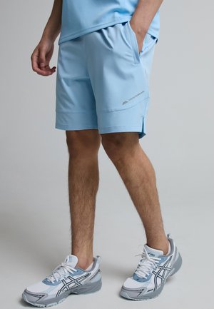 Person iført lyseblå sportsshorts med "MONTERRAIN" logo og hvide-grå Asics sneakers, stående med den ene hånd i lommen.