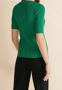 Kvinna som bär en kortärmad, ribbad grön topp och svarta byxor, stående med ryggen delvis vänd mot en beige bakgrund.