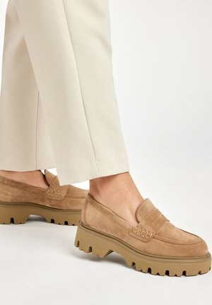 Mocassini in suede beige con una suola in gomma spessa e testurizzata che presenta un bordo frastagliato. Il design include un dettaglio cucito sul davanti.