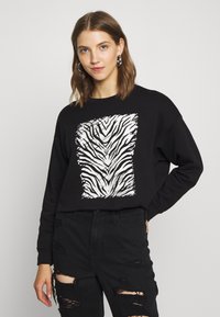 Sweatshirt preto cropped com gráfico de riscas de zebra brancas. Apresenta um decote redondo e mangas longas, combinado com jeans pretos desgastados.