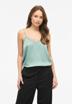 Femme aux longs cheveux bruns portant un caraco vert clair orné de dentelle et un pantalon noir, debout sur un fond blanc uni.