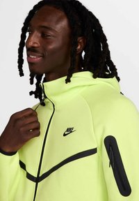 La sudadera con cremallera de Nike en amarillo neón presenta detalles en negro, un bolsillo con cremallera en la manga y una tela suave y texturizada.