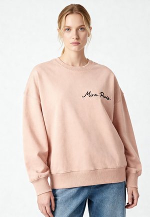 Jeune femme aux cheveux blonds portant un sweat-shirt ample rose clair avec le texte « Mira Paris » et un jean bleu sur fond blanc.