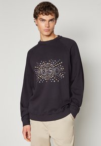 Sudadera negra con mangas raglán, que presenta un patrón decorativo de tachuelas metálicas y el logo "Hugo" en el centro.