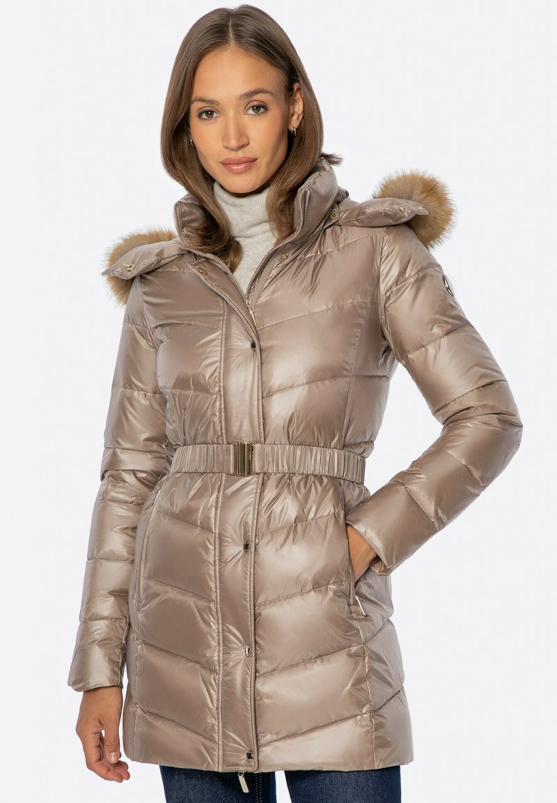 Mujer con un abrigo acolchado brillante taupe ceñido con cinturón, capucha con ribete de piel y suéter de cuello alto beige, de pie con una mano en el bolsillo.