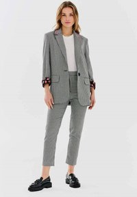 Szary żakiet w kratę houndstooth z klapkami i haftowanymi mankietami, w połączeniu z dopasowanymi spodniami i czarnymi loafersami. Gładka tekstura, strukturalny krój.