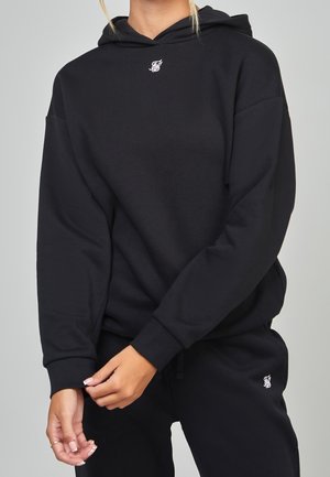 Osoba ubrana w czarną bluzę z kapturem i dopasowane czarne spodnie dresowe, regulująca rękaw obiema rękami, na jednolitym jasnym tle.