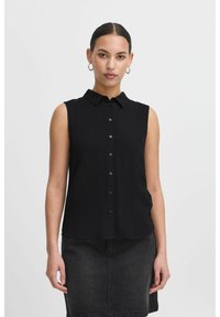 Maglietta nera senza maniche con colletto, chiusura frontale con bottoni e texture morbida. Abbinata a una gonna di jeans nera.