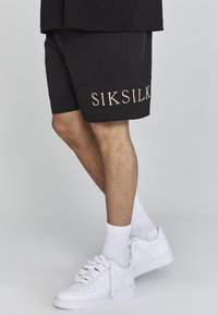 Black shorts with an embroidered "SIKSILK" logo in gold, paired with white sneakers and white crew socks, showcasing a simple athletic style.