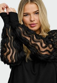 Svart overdel med lange, transparente ermer med teksturerte volanger. Kroppen er ensfarget svart med en avslappet passform og ribbestrikkede mansjetter.