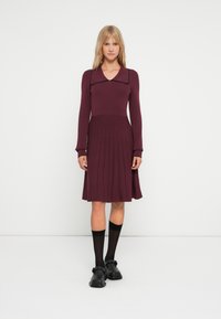 Abito in maglia bordeaux con colletto, maniche lunghe, gonna plissettata e colore uniforme. Abbinato a calzini e scarpe nere.