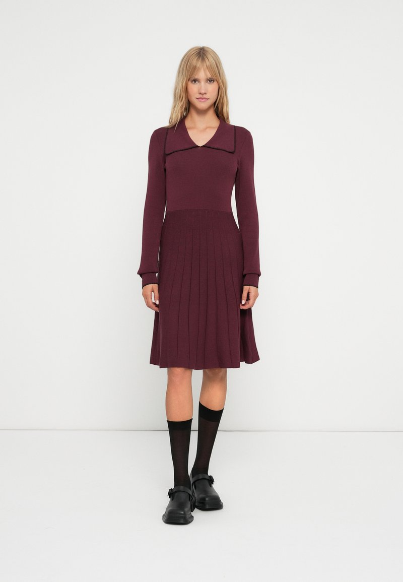 Abito in maglia bordeaux con colletto, maniche lunghe, gonna plissettata e colore uniforme. Abbinato a calzini e scarpe nere.