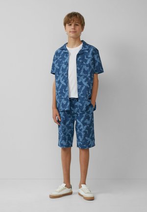 Camisa de botón de manga corta y pantalones cortos a juego en un estampado de hojas tropicales azules, combinados con zapatillas blancas y una camiseta blanca lisa.
