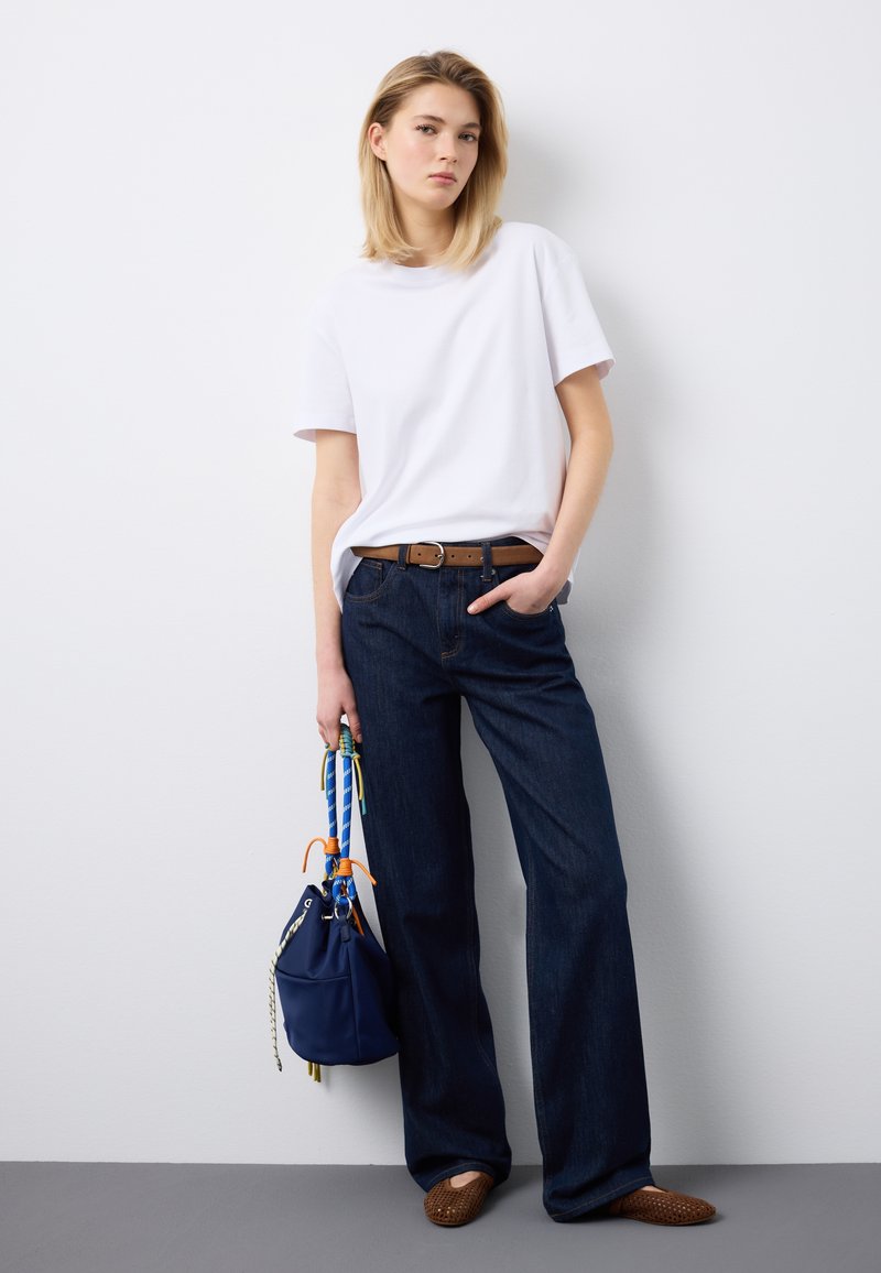 Frau steht an der Wand, trägt ein weißes T-Shirt, dunkelblaue Jeans, einen braunen Gürtel und braune Schuhe, hält eine marineblaue Handtasche mit bunten Trägern.