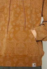 Hombre con sudadera con capucha de patrón marrón, con la mano en el bolsillo delantero, mostrando la etiqueta de la marca Carhartt en el dobladillo acanalado.