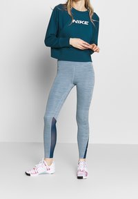 Sweat-shirt court de couleur sarcelle avec logo "NIKE" blanc, assorti à un legging gris et bleu avec des accents en mesh. Chaussures de sport blanches et roses.