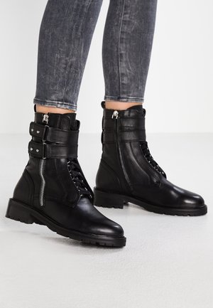 Personne portant des bottines en cuir noir avec zips latéraux et double lanière à boucle, assorties à un jean skinny noir délavé.