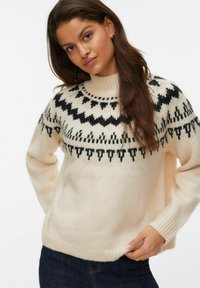 Frau mit langen braunen Haaren, die einen cremigen Pullover mit schwarzen geometrischen Mustern über der Brust und den Schultern trägt, kombiniert mit dunklen Jeans.
