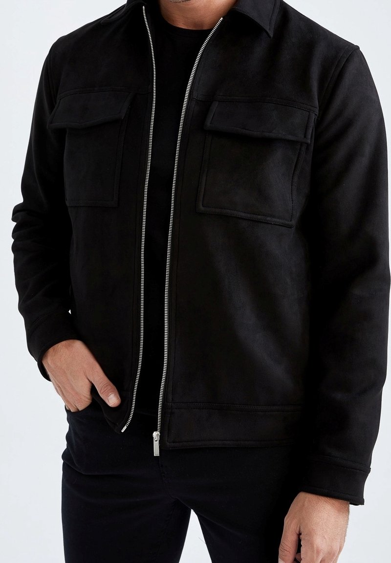 Giacca in suede nera con zip sul davanti, due tasche sul petto e collo. Il tessuto ha una texture liscia e una finitura opaca.