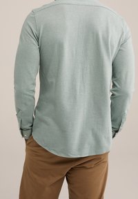 Man i ljusgrön långärmad skjorta och bruna byxor, står med händerna i fickorna, vänd bort från kameran mot en enfärgad bakgrund.