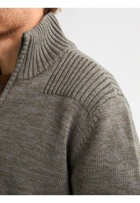 Strickpullover in Olivgrün mit hohem Rippkragen und strukturierten Schulterdetails. Das Material wirkt weich und warm mit einem dezenten Muster.