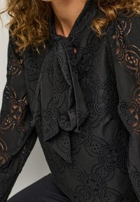 Närbild av en person som bär en svart spetsblus med blommönster och en knuten rosett vid halsen.