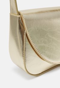 Detailní záběr na kovově zlatou koženou kabelku přes rameno s klopou a viditelným prošíváním podél okrajů.