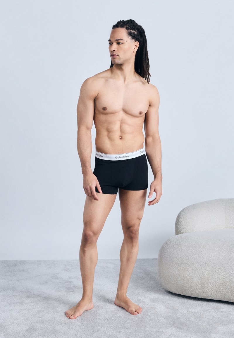 Lihaksikas mies, jolla on rastat ja joka on pukeutunut mustiin Calvin Kleinin bokserishortseihin, seisoo paljain jaloin maton päällä valkoisen pyöreän tuolin lähellä.