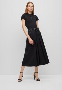 Schwarzes Poloshirt mit kurzen Ärmeln, kombiniert mit einem hoch taillierten schwarzen Rock mit Falten und einem breiten Gürtel mit einer Schnalle. Schwarze High-Heels Sandalen.