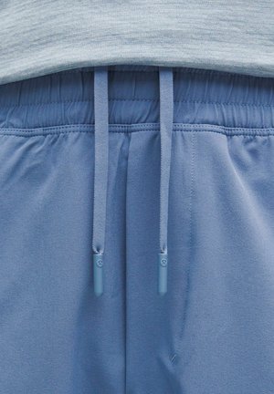 Pantaloni sportivi blu con una texture liscia, dotati di chiusura con lacci e vita elastica. I dettagli includono tappi agli estremi dei lacci.