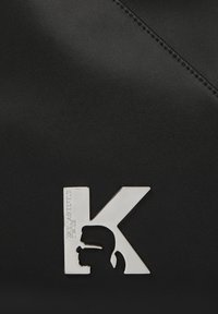 Chrome metallic "K" emblem with a cutout silhouette of a man's head and "KARL LAGERFELD JEANS" text on black leather background.