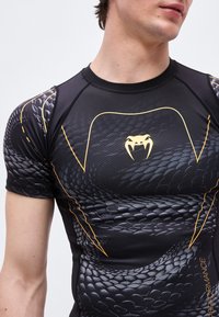 Jonge man die een zwart sportshirt draagt met een slangenhuidpatroon en een geel slangentandenlogo op de borst.