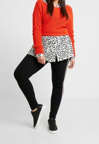 Maglione rosso a righe sovrapposto a una camicia a stampa leopardata bianca e nera, abbinato a leggings neri con una striscia laterale e sneakers nere.