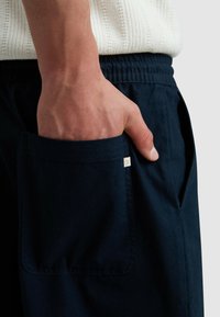 Navyblauwe broek van zacht materiaal, met een zijzak en een klein label. De tailleband is elastisch voor comfort en flexibiliteit.