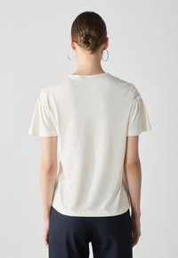 T-shirt a maniche corte color crema con dettaglio arricciato sulle spalle, scollatura a girocollo e materiale in cotone morbido, visto da dietro.
