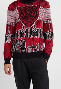 Roter Strickpullover mit einem auffälligen schwarzen Bär-Motiv, Stadtsilhouette und geometrischen Mustern in Schwarz-Weiß-Details.