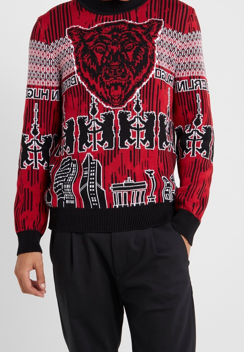 Roter Strickpullover mit einem auffälligen schwarzen Bär-Motiv, Stadtsilhouette und geometrischen Mustern in Schwarz-Weiß-Details.