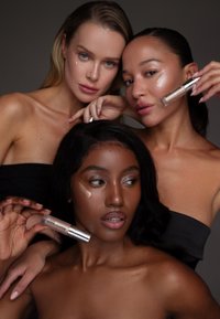 Drei Personen präsentieren einen flüssigen Highlighter in silberner Verpackung. Ihre Hauttöne variieren und haben einen glatten, reflektierenden Glanz. Sie tragen schwarze und neutrale Outfits.