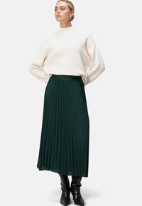 Jupe maxi plissée verte avec une texture lisse, associée à un col roulé blanc doux à côtes et des bottes noires arrivant au genou.