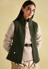 Grüne Fleece-Weste mit hohem Kragen, Reißverschluss vorne und Seitentaschen, kombiniert mit einem cremefarbenen, gerippten Pullover und einem gemusterten Hemd.