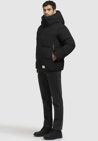 Chaqueta acolchada negra con cuello alto y capucha ajustable, que cuenta con bolsillos con cremallera y un parche rectangular con el logo en el dobladillo.