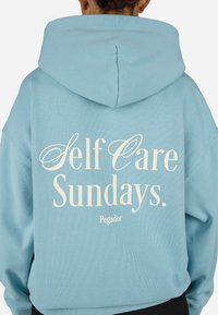 Ανοιχτό μπλε φούτερ με μαλακή υφή. Διαθέτει λευκό εκτυπωμένο κείμενο "Self Care Sundays." και "Pegador" στην πλάτη. Υπάρχει κουκούλα.